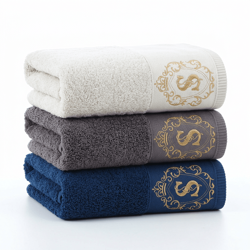 Luxury hotel bath towel in white with gold embroidered crest on a white background.